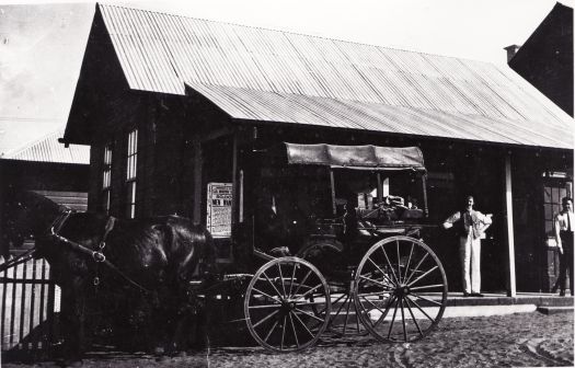 Canberra's first Post Office at Acton showing two men standing in front of a weatherboard building and a horse drawn carriage. Also visible is a recruiting poster for men to serve in World War 1. The men are believed to be George Bondfield, the postmaster, and Bill Christie the telegrapher who were both appointed in 1914.