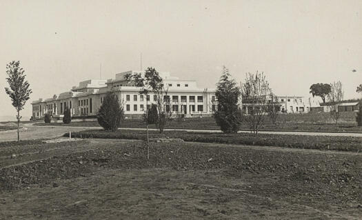A right side front view of Parliament House within a few years of its construction. The surrounding trees and shrubs are small in size and bare patches of dirt are obvious.