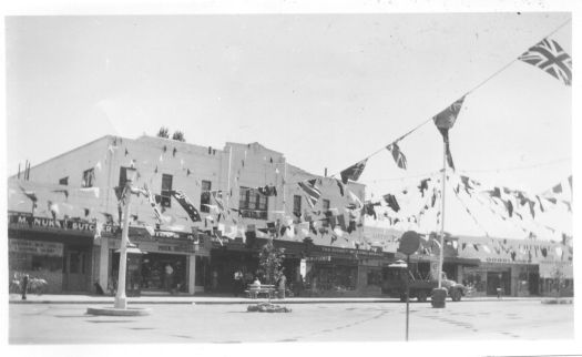 View of Franklin Street showing the shops - butchers, chemist and newsagency - with bunting and flags strung across the road.
