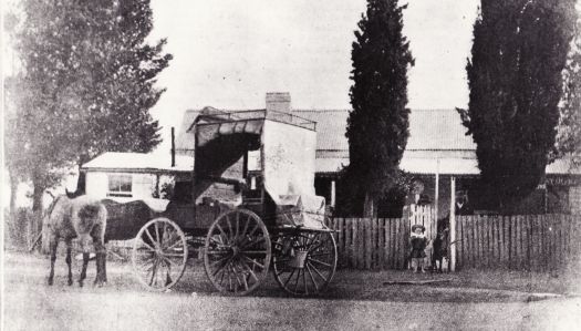 Arrival of the mail at Ainslie post office. Shows two horses and a four wheeled enclosed wagon.  Two small children are standing at the gate in front of the post office.