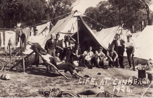 Workmens camp, showing eleven unidentified men of various ages, some sitting and some standing, with tents in the background and washing hanging from lines. Two men are holding banjos and another an accordion.