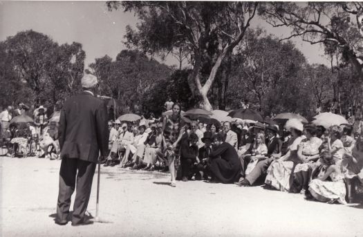 President of the CDHS, Cam Morris, addressing assembly at Commencement Stone ceremony on Canberra Day. The photograph shows the Commencement Stone site in bushland on Capital Hill.  Most of the guests, including the Minister for the Capital Territiry, Tony Staley, are seated. Actors, dressed in period costume for a re-enactment of the naming ceremony in 1913, are in the front row.