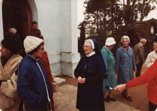 Outside old Catholic Church, Meehan Street, Yass. Shows several people,  including a nun, in coats outside the church and a grotto at right