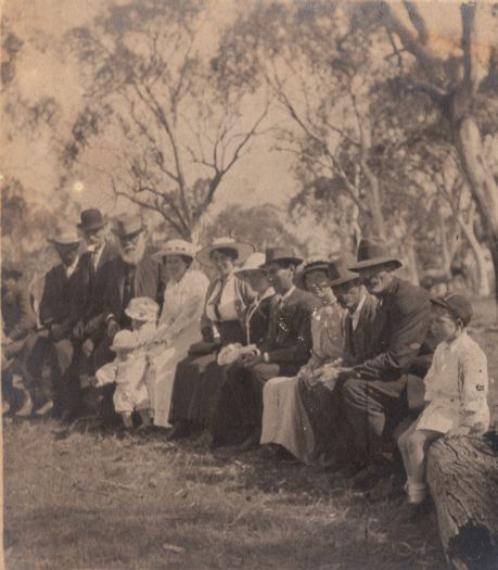 Noted as Thompsons and James (Joe), Nimmitabel. Shows a group of ten adults and three children, dressed in their finest clothes, sitting on a log possibly at a race meeting at Nimmitabel to the east of Cooma.