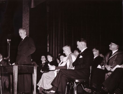 Conferring of ANU degrees at Albert Hall. Ben Chifley speaking, Mrs Chifley, Sir George Currie and others sitting.