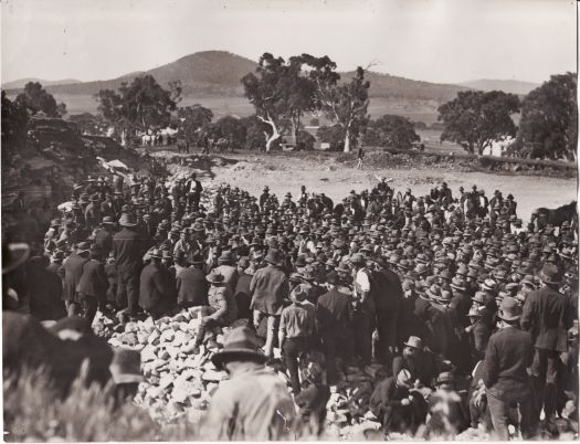 Photo shows several hundred workmen, most in hats and coats, assembled in quarry on Capital Hill. Mount Ainslie and possibly East Block are visible in the background.