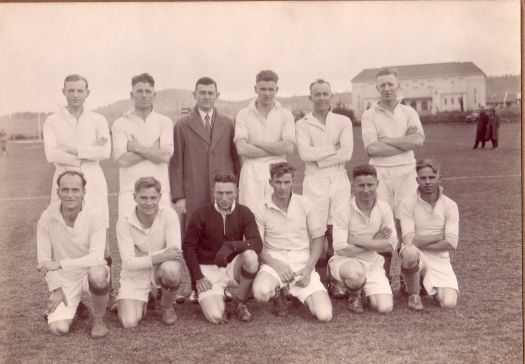 Group of twelve men showing the Capitol Theatre, Manuka in the background. Eleven of the men are dressed for a football match, one like a soccer goalkeeper. The other man is C.S. Daley who is standing in the back row in coat and tie.