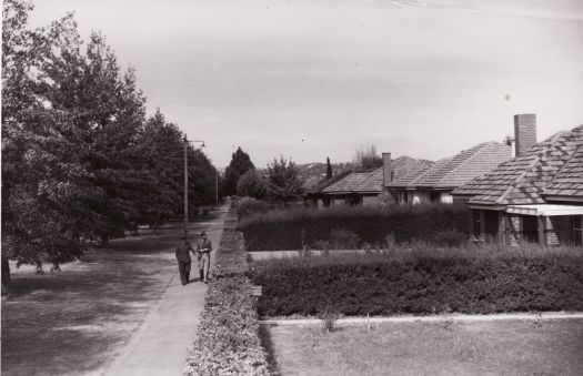 Shows two men walking down a suburban street with trees and hedges between the houses. Possibly Donaldson Street, Braddon.
