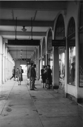 Photo shows people walking and standing on a colonnaded footpath - probably the Sydney Building - along Northbourne Avenue. Signs for a chemist and the 'Capitol Cafe' are visible.