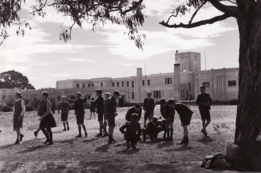 Canberra High School. Children are in the playground and some boys are playing marbles while some girls walk by.
