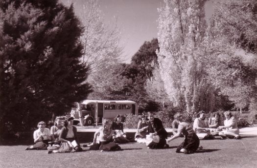 Group of women sitting in the sun on the lawns in front of Albert Hall, bus in the background.