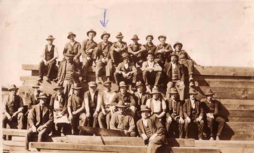 Group photo of a construction gang, who were building the Hotel Canberra, sitting on a stack of timber.