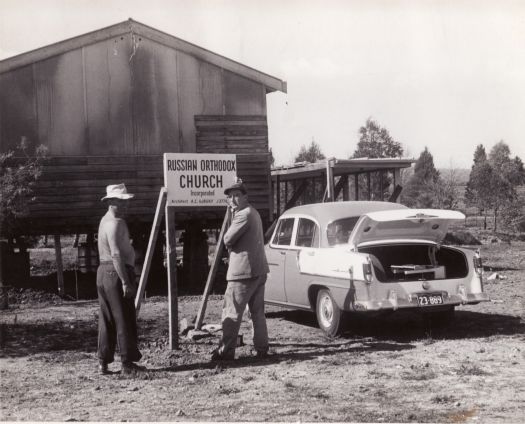 Two men in front of sign for Russian Orthodox Church with temporary building behind. Holden car parked in front