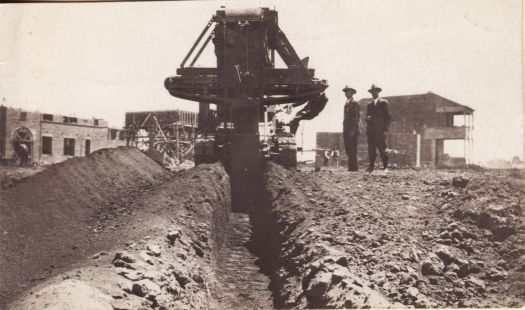 Trench digger at work in Civic. Two suited men are standing nearby.