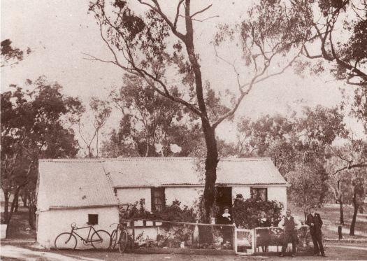 Family portrait in front of the house erected at Duntroon by Frank Dowthwaite. A eucalypt stands in the middle of the garden and two bicycles are leaning against the house and fence. The house is surrounded by woodland.