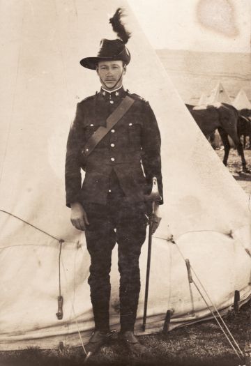 Light horseman standing in front of a bell-shaped tent. Subject possibly is an officer as he has a sword. The subject is standing on grass and the hindquarters of horses in the middle foreground are also on grass.