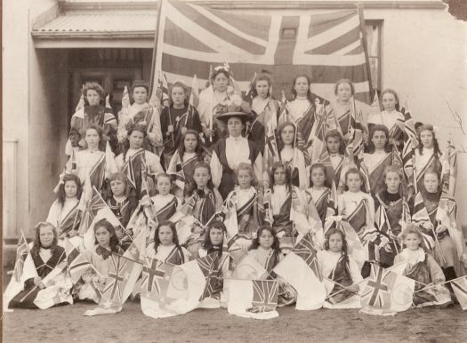 Group of 32 girls and one female (possibly their teacher) outside a building and in front of a large Union Jack. Each girl is holding a flag.