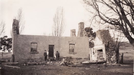 Roofless house made of pise with two men standing in front of it. 