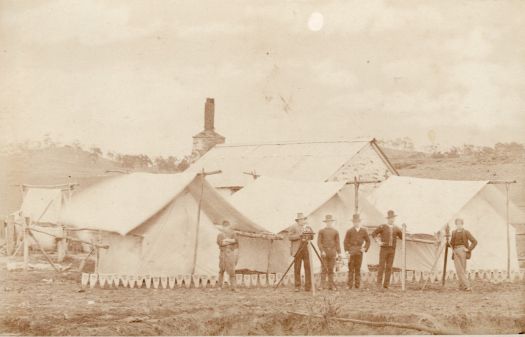 Surveyor campsite, FCT. Shows six men standing in front of tents with a house in the background. One of them is standing behind a theodolite.