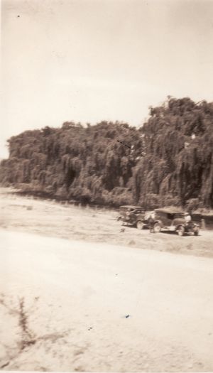 Camp at the "Willows" about 12 miles from Cooma on the Queanbeyan road. Two vehicles are stopped near a river with three people nearby.