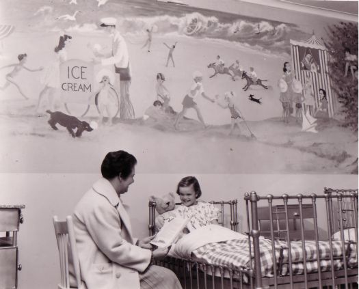 Children's ward, Canberra Hospital. Woman reading to a young girl sitting up in a hospital bed. She is holding a teddy bear. A mural has been painted on the wall behind her.