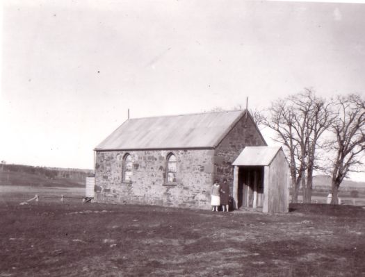 St Ninian's Church, Brigalow St, Lyneham. Photograph taken before suburban development from south east of the church looking in the direction of Yass Road (now Mouat Street). Taken before second extension, shows two women standing near the front entrance and the two elm trees (planted c1873, removed in 2001) are at the back (without leaves, suggesting winter).