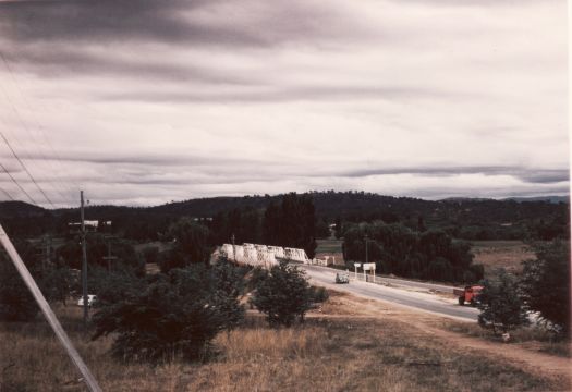 Commonwealth Avenue bridge taken from the vicinity of Cathedral Hill showing cars crossing the old wooden truss bridge and trees lining the banks of the Molonglo River