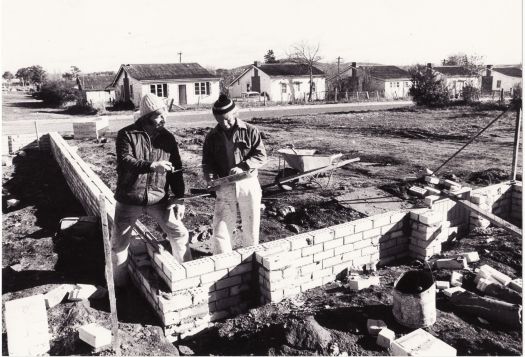 Replacement houses under construction at the Causeway in Kingston. The photo shows two bricklayers building walls. Timber houses are in the background.