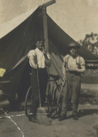 Joe Harrington and F.L. McDonald are standing in front of a tent, smoking cigarettes. A bicycle is leaning against a tent pole.  One of the men is holding a broom and the other a shovel.