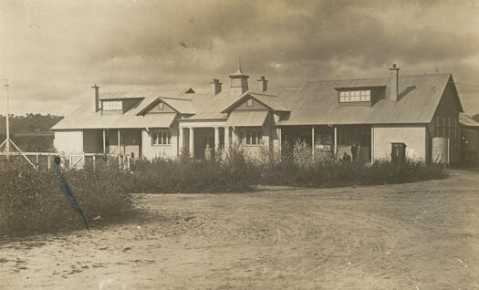 Photograph shows a long wooden building with four chimneys. Two men, one sitting and the other standing, are on the porch at one end of the building.