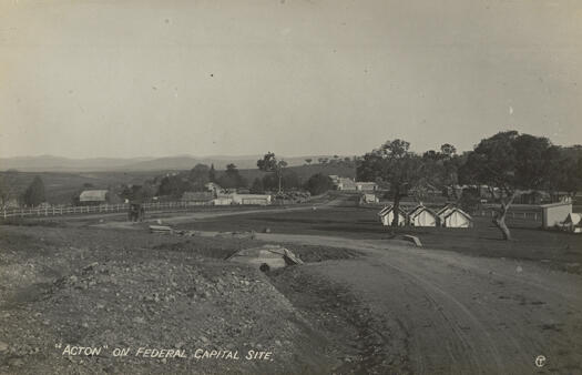 Postcard shows a road leading leading to a building in the background towards Lennox Crossing. The road has a post and rail fence along its boundary. A horse and carriage are in the middle foreground and a covered haystack in the background. In the foreground are eight tents and a building.