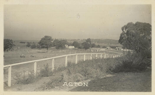 The postcard shows a white fence in the foreground with a building in the middle distance.