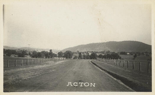 Taken from the middle of the road leading inot Acton. Fences are on both sides of the road with building and trees in the middle distance.