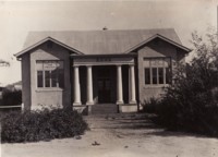 The first branch of the Commonwealth Bank at Acton. The photograph shows a front and side view of the bank building and a fence running along te side of the building.