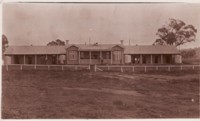 Bachelors quarters at Acton. Front view shows the entrance with a white rail fronting a verandah. Four water tanks are next to the building.