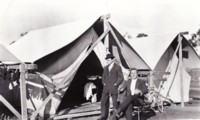 D.P. Russell is sitting on a chair and Charles Laverty is standing in front of their temporary home, a tent at Acton. Both men appear dressed in their best clothes. A bicycle is shown behind Russell and Laverty is wearing a hat. A washstand with a dish is visible just inside the tent and a water jug is sitting on the duck boards.
