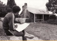 A young man is sitting on a stone wall sketching Blundell's Cottage. The cottage is seen from a close view with a white paling fence running off to the left.