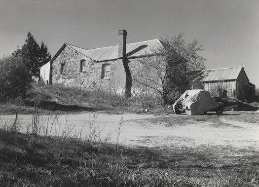 Side view of Blundell's Cottage. A road runs from left to right and a car is parked on the road covered by a torn cover. The number plate is 27473. A shed is on the right side of the cottage.