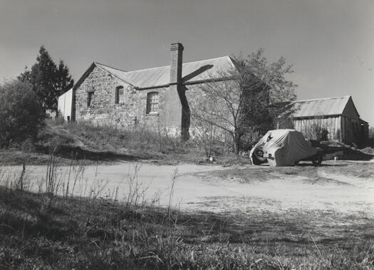 Side view of Blundell's Cottage. A road runs from left to right and a car is parked on the road covered by a torn cover. The number plate is 27473. A shed is on the right side of the cottage. See copy of part of this photo in NCDC Annual Report 1961-62