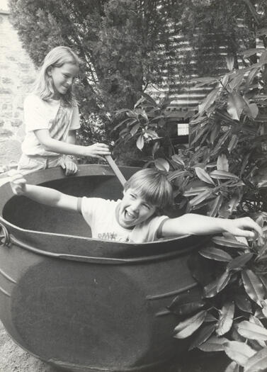 A young boy, in a cauldron, is being poked by a young girl. A water tank on a tank stand, is in the background with shrubs surrounding the tank stand.