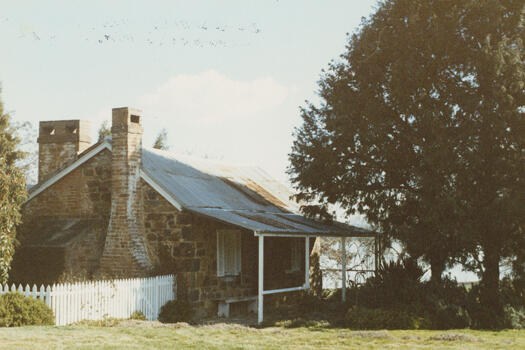 Side view of Blundell's Cottagefrom Wendouree Drive. Lake Burley Griffin can be seen through a large tree on the right.