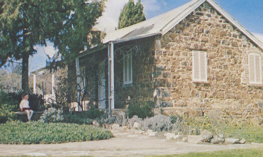 Photograph shows the southern side and front of Blundell's Cottage. A small child is sitting on a seat in fron of the cottage.