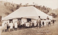 Cotter Junction Public School showing a tent with 24 students of different ages lined up in front. Also shows two males, probably teachers, each wearing a suit and tie. The man standing in the doorway is the teacher, Sidney Marshall.