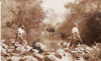 Two unidentified men crossing the Cotter River. One man is carrying a rifle in his right hand. The other man is poking a stick into the water.