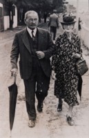 Arthur and Celia Wilden, of Queanbeyan, walking along a street. Celia is carrying gloves and umbrella, wearing a hat and carrying a handbag over her left arm. Arthur is wearing a suit with tie and waistcoat and is also carrying an umbrella.