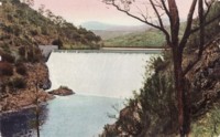 Front view of the Cotter Dam overflowing. Shows steps leading up to the dam wall on the right side of the photo. People (ant-like in size) can be seen on the viewing platform.