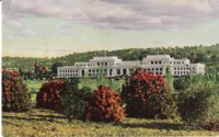 Front view of Parliament House. Text on back of postcard: Parliament House, Canberra, seat of the Australian Government; opened May 9th 1927 by H.R.H. the Duke of York. The cost about 700,000 pounds; covers 4 acres with 168 acres of surrounding gardens. Church of St. Andrew in rear at left. Red Hill is showing in the background