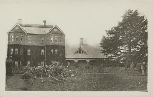 Front view of Government House, Yarralumla showing a 3 story building with a single story building, the original residence, attached. The photo shows part of the garden with two men standing to the right.