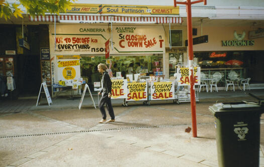 Front of Develin's Chemist's in Garema Place, Civic. There are four  sandwich boards on the footpath stating there is a closing down sale. There is a young person walking past the front of the shop.