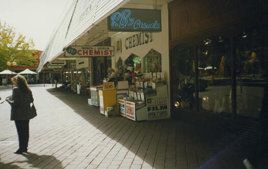 View along the footpath at Garema Place. Shows Patrick J. Develin's Chemist; R&B's casual shop is closest to the camera. A woman is standing with her back to the camera.